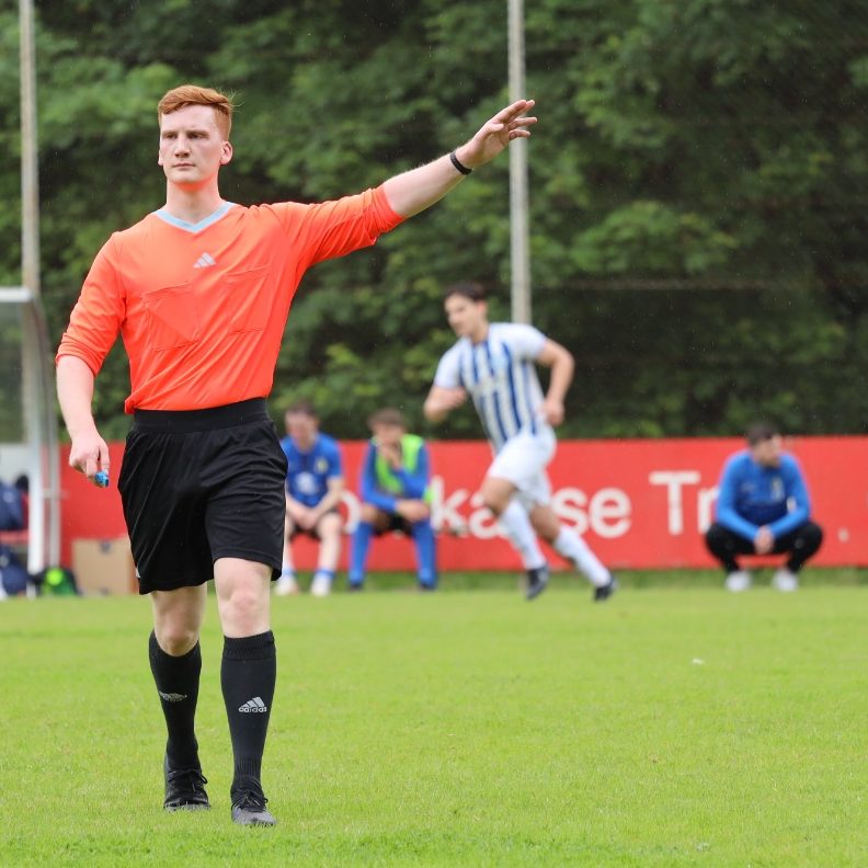 Jonas Klar als Fußballschiedsrichter – entschlossene Spielleitung im roten Schiedsrichtertrikot, Spieler im Hintergrund